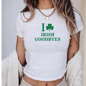 May include: White t-shirt with the text "I Irish Goodbyes" in green, featuring a green shamrock. The shirt has short sleeves and a crew neck. A gold chain necklace is visible.