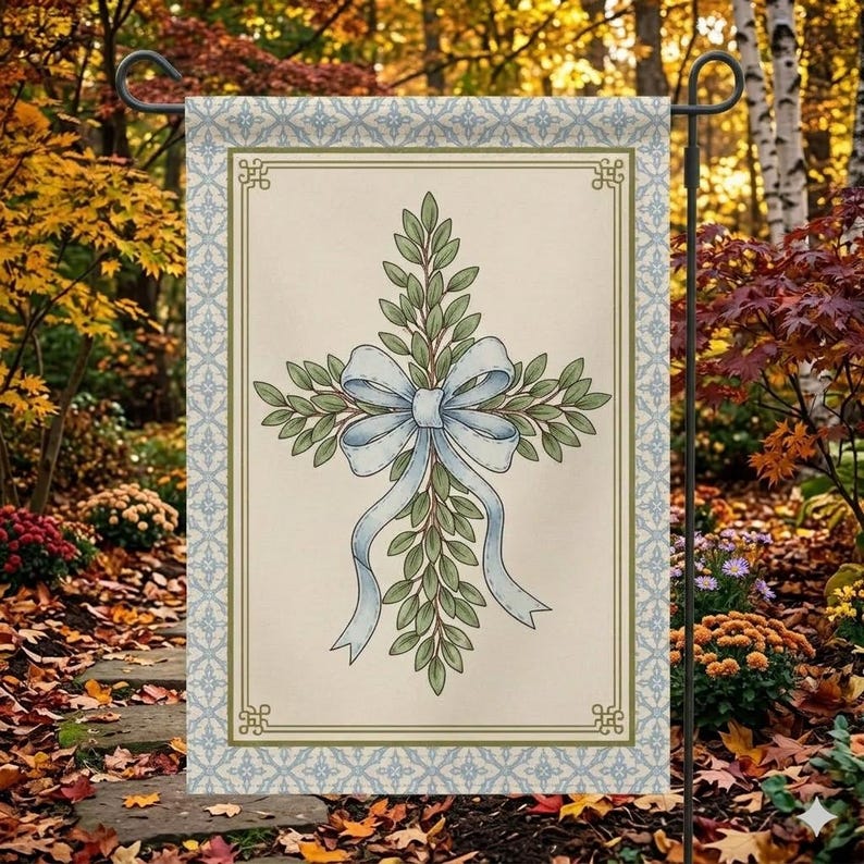 May include: A garden flag featuring a cross design made of green leaves and a light blue bow. The flag has a cream-colored background with a decorative border in blue and green. The flag is displayed outdoors.