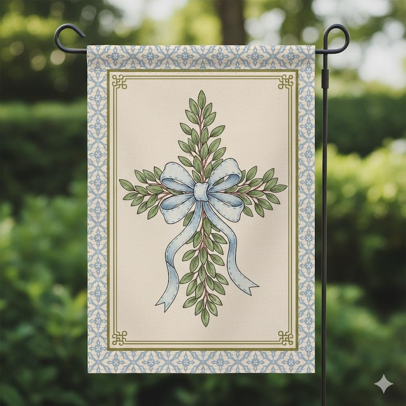 May include: A garden flag featuring a cross design made of green leaves and a light blue bow. The cross is centered on a cream-colored background, framed by a blue and cream patterned border. The flag is displayed on a black metal stand.