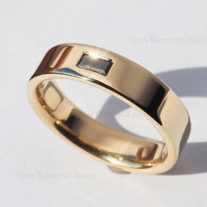 Puede incluir: Un anillo dorado con una piedra rectangular de color oscuro engastada en el centro. El anillo tiene una superficie lisa y pulida y un diseño ligeramente curvado. El anillo se muestra sobre una superficie blanca.