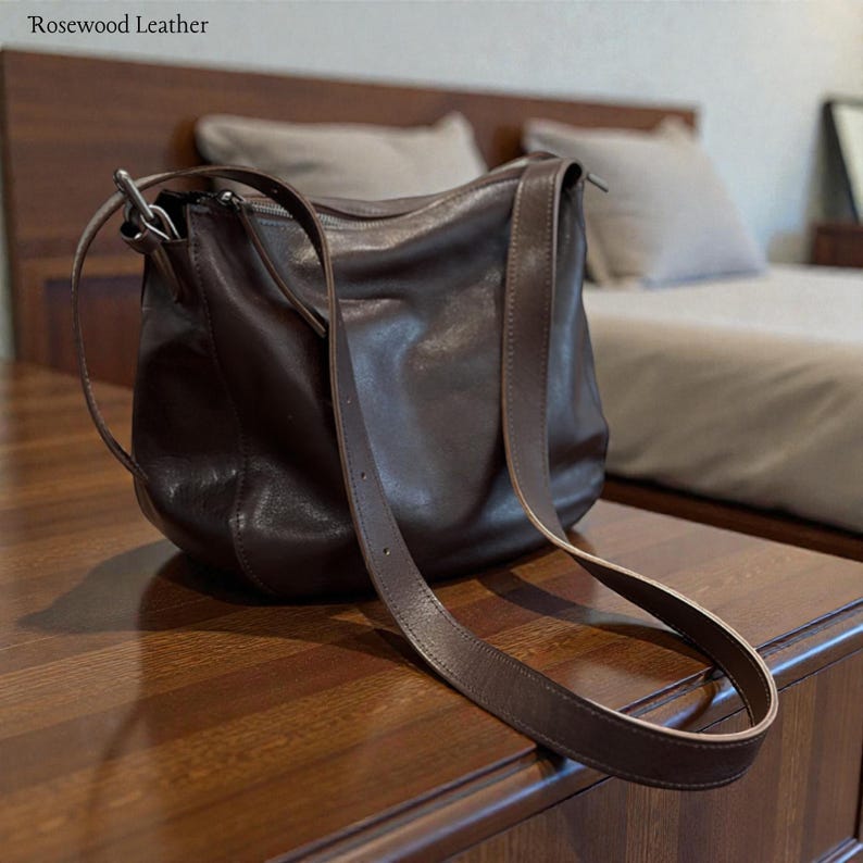 May include: A dark brown leather shoulder bag with a long strap. The bag has a rounded shape and a zipper closure. The text "Rosewood Leather" is in the top left corner.