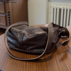 May include: A dark brown leather shoulder bag with a long strap. The bag is sitting on a wooden table. The text "Rosewood Leather" is in the top left corner.