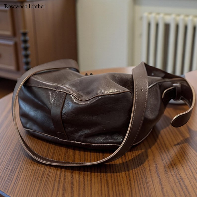 May include: A dark brown leather shoulder bag with a long strap. The bag is sitting on a wooden table. The text "Rosewood Leather" is in the top left corner.