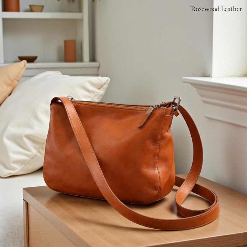 May include: A brown leather shoulder bag with a long strap. The bag has a zipper closure and is sitting on a wooden surface. The text "Rosewood Leather" is visible in the upper right corner.