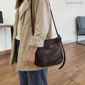 May include: A dark brown leather crossbody bag with a long strap. The bag is held by a person wearing a tan jacket and blue jeans. The text "Rosewood Leather" is in the upper right corner.