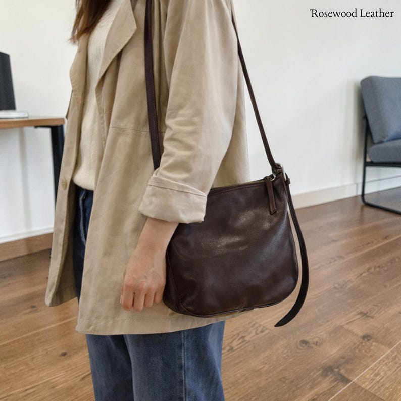 May include: A dark brown leather crossbody bag with a long strap. The bag is held by a person wearing a tan jacket and blue jeans. The text "Rosewood Leather" is in the upper right corner.