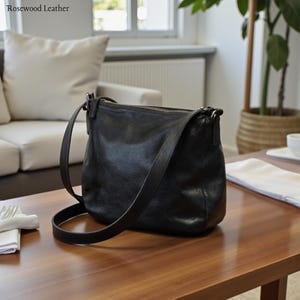 May include: A black leather crossbody bag with a long adjustable strap. The bag has a zipper closure and a slightly textured surface. The bag is sitting on a wooden table. The words "Rosewood Leather" are in the top left corner.
