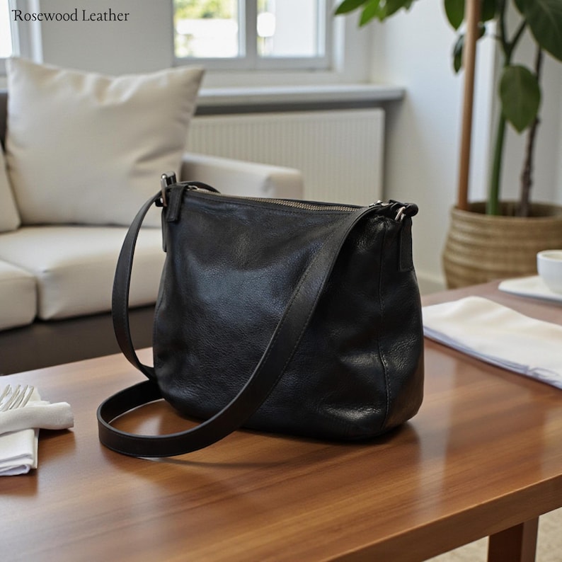 May include: A black leather crossbody bag with a long adjustable strap. The bag has a zipper closure and a slightly textured surface. The bag is sitting on a wooden table. The words "Rosewood Leather" are in the top left corner.