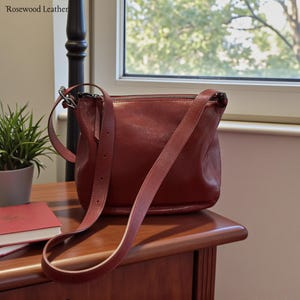 May include: A burgundy leather crossbody bag with a long adjustable strap. The bag has a zipper closure and is sitting on a wooden surface. A small potted plant and a book are also visible.