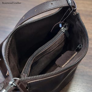 May include: A brown leather shoulder bag with a zipper closure. The bag has a long strap and an interior pocket with a zipper. The bag is open, revealing the interior lining. The text "Rosewood Leather" is in the top left corner.