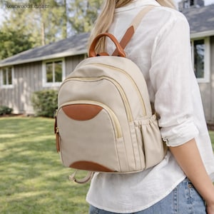 May include: A beige nylon backpack featuring brown leather accents, a top handle, and adjustable shoulder straps. The bag includes a main zippered compartment, a smaller front zippered pocket with a leather patch, and side pockets with elasticated edges.