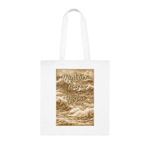 May include: White tote bag with a sepia-toned image of ocean waves. The text "Mightier than the waves of the sea is His love for you" is printed on the image. The bag has two white handles.