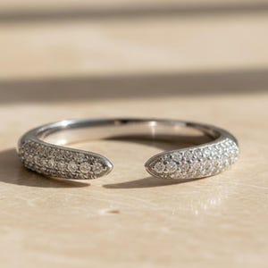 May include: A silver ring with a split design, featuring a band encrusted with small, sparkling gemstones. The ring has an open front, creating a unique and modern look. The gemstones catch the light, adding brilliance.