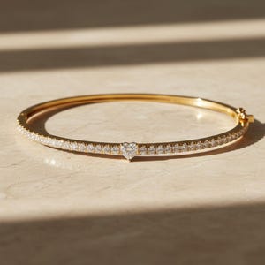 May include: A gold bangle bracelet with a heart-shaped diamond in the center. The bracelet is encrusted with small, round diamonds along the front half. The bracelet is displayed on a light-colored surface.
