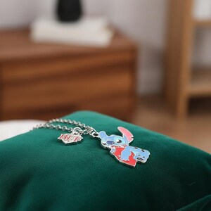 May include: A silver necklace featuring a blue, red, and white enamel pendant of the character Stitch from Lilo & Stitch. The necklace also includes a small BFF charm. The pendant is displayed on a green velvet surface.