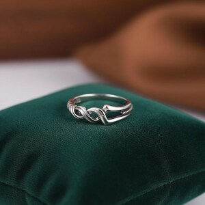 May include: A silver ring with an infinity symbol design. The ring is displayed on a dark green velvet surface. The ring has a polished finish and a delicate, intricate design, perfect for everyday wear.