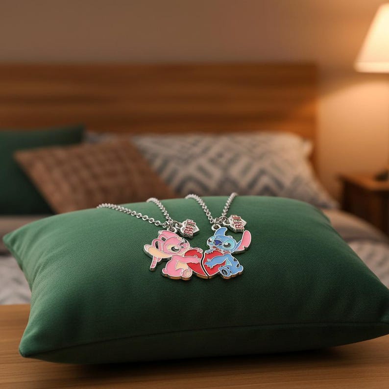 May include: Two matching necklaces with cartoon character pendants, one pink and one blue, each holding a heart. The pendants are on silver chains, resting on a green pillow. The background includes a bed and a lamp.