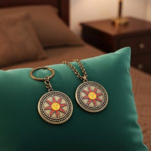 May include: A bronze pendant necklace and keyring featuring a sun design with red and yellow detailing. The sun has a face at its centre and is encircled by a dotted border. The items are displayed on a green cushion.