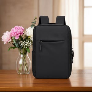Könnte beinhalten: Ein schwarzer Rucksack mit rechteckigem Design. Der Rucksack hat eine Vordertasche mit Reißverschluss und eine Seitentasche. Die Träger sind schwarz und gepolstert. Links vom Rucksack steht eine Vase mit Blumen.