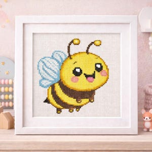 May include: A framed cross-stitch of a cartoon bee with a yellow body, brown stripes, and blue wings. The bee has a smiling face with a pink tongue sticking out. The artwork is displayed in a white frame.