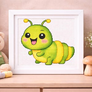 May include: A framed cross-stitch of a cartoon caterpillar. The caterpillar is green and yellow with a smiling face and pink cheeks. The artwork is displayed in a white frame, and is on a wooden surface.