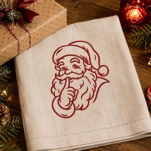 May include: A folded off-white linen napkin featuring a red embroidered line art illustration of Santa Claus with a finger held to his lips in a shushing gesture. The design shows Santa wearing a hat and sporting a full beard and moustache.