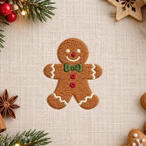 May include: An embroidered gingerbread person patch featuring a brown textured body, a green bow tie, two pink buttons, a red nose, and white wavy icing details on the arms and legs, centered on a light beige fabric background.