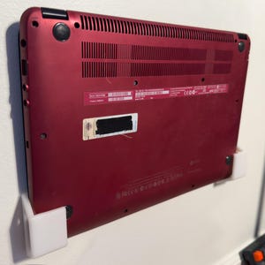 Op de afbeelding: Een rode laptop, van onderaf gezien, gemonteerd op een witte wandbeugel. De laptop heeft een reeks ventilatieopeningen en poorten, en een sticker met onduidelijke tekst. De laptop is gemonteerd op een witte wandbeugel.