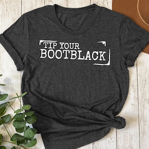 May include: Dark gray t-shirt with the text "TIP YOUR BOOTBLACK" in white block letters. The shirt is knotted at the bottom and has rolled-up sleeves. A brown hat and green plant are in the background.
