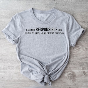 May include: A light gray t-shirt with the text "I AM NOT RESPONSIBLE FOR THE WAY MY FACE REACTS WHEN YOU SPEAK" in black. The shirt is knotted at the bottom and has rolled-up sleeves. The shirt is displayed on a light wood-grain surface.