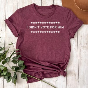 May include: Maroon t-shirt with the text "I DIDN'T VOTE FOR HIM" in white, with star accents above and below the text. The shirt is folded and knotted at the bottom. A brown hat and green plant are in the background.