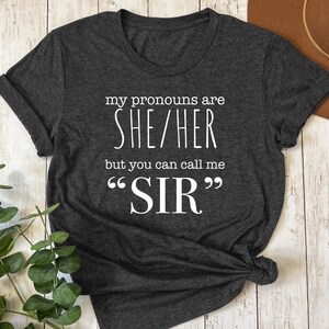 May include: Dark gray t-shirt with the text "my pronouns are SHE/HER but you can call me SIR" in white. The shirt is knotted at the bottom and has rolled-up sleeves. The background includes a wooden surface and greenery.