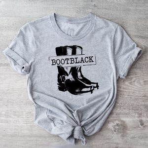 May include: A light gray t-shirt with a black graphic of a cowboy boot and the word "BOOTBLACK" in a rectangular box. The shirt is knotted at the bottom.