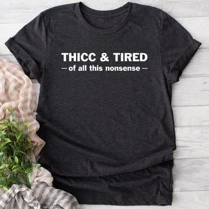 May include: Dark gray t-shirt with the text "THICC & TIRED - of all this nonsense -" in white. The shirt is folded, with a plant and a patterned scarf in the background. The shirt is made of a soft material.