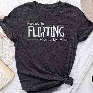 May include: Dark gray t-shirt with the text "oblivious to FLIRTING please be blunt" in white. The shirt is knotted at the bottom. A hat, candle holder, book, and coffee cup are also in the image.