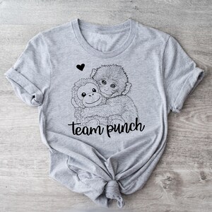 May include: A light gray t-shirt featuring a black and white illustration of two monkeys hugging, with a small black heart above them. The text "team punch" is written in a script font below the monkeys.