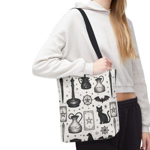 May include: White tote bag with a black strap featuring a black and white illustration of various mystical objects. The design includes a black cat, potion bottles, tarot cards, a bat, and a witch's hat.
