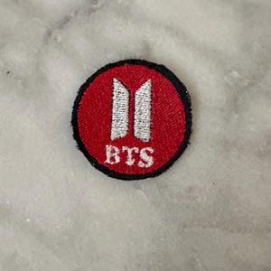 以下が含まれることがあります： 赤色の背景に白いデザインと白い文字「BTS」が施された、丸い刺繍ワッペン。 パッチには黒い縁取りがあります。 おそらくロゴまたはエンブレムです。