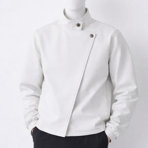 Puede incluir: Una chaqueta blanca con cuello alto y cierre frontal asimétrico con dos botones de color marrón oscuro. La chaqueta tiene mangas largas con puños acanalados y se muestra sobre un pantalón negro liso en un maniquí.
