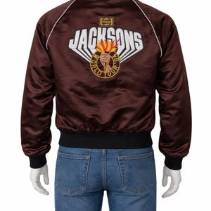 Könnte beinhalten: Braune Satin-Bomberjacke mit dem weißen Schriftzug "JACKSONS" und einem Logo mit einer Fackel und den Worten "WORLD TOUR 84-85". Die Jacke hat schwarze Bündchen, Kragen und Saum sowie weiße Paspeln.