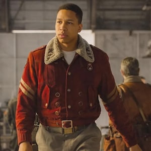 May include: A burgundy bomber jacket with a cream-colored shearling collar. The jacket features gold stripes on the sleeves, a matching belt, and a button-up front. The jacket is worn over a white shirt and paired with gray pants.