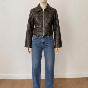 May include: A brown faux leather jacket with a snakeskin pattern, paired with blue jeans and black pointed-toe boots. The jacket has a collar and a zipper closure. The jeans are straight-legged.