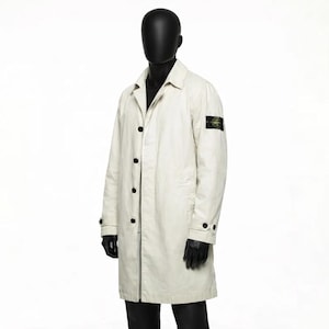 May include: A cream-colored, button-down coat with a classic collar and long sleeves. The coat features black buttons and a small, square patch on the upper left sleeve. The coat is displayed on a black mannequin.