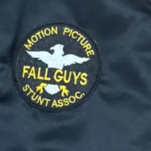 Pode incluir: Casaco preto com um patch circular. O patch diz "MOTION PICTURE" e "STUNT ASSOC." em letras amarelas. As palavras "FALL GUYS" e um emblema de &aacute;guia branca tamb&eacute;m est&atilde;o presentes. O casaco &eacute; feito de um material escuro e liso.