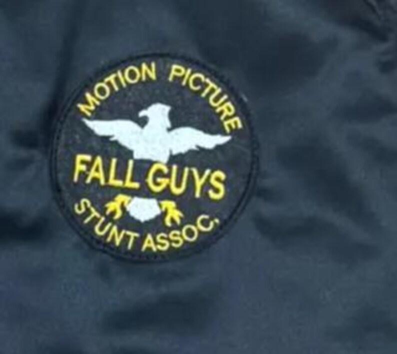 Pode incluir: Casaco preto com um patch circular. O patch diz "MOTION PICTURE" e "STUNT ASSOC." em letras amarelas. As palavras "FALL GUYS" e um emblema de &aacute;guia branca tamb&eacute;m est&atilde;o presentes. O casaco &eacute; feito de um material escuro e liso.