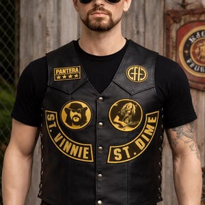 Op de afbeelding: Zwart leren vest met goud- en zwarte patches. Het vest heeft de bandnaam "Pantera" op de voorkant en een "St. Vinnie St. Dime" ontwerp. Een zwarte baseball cap met het "Pantera" logo is ook zichtbaar.