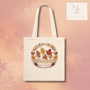 Könnte beinhalten: Eine cremefarbene Stofftasche mit einer kreisförmigen Herbstillustration von fünf anthropomorphen Blättern, die Mützen und Schals tragen. Der Text WENN DIE BLÄTTER TANZEN BEGINNT DIE SCHÖNSTE JAHRESZEIT ist in einem Kranz aus Blättern abgebildet.
