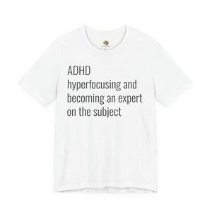 Op de afbeelding: Een wit T-shirt met korte mouwen en zwarte tekst: ADHD hyperfocusing and becoming an expert on the subject. Het T-shirt heeft een ronde hals en een klein, kleurrijk hersenlogo aan de binnenkant van de kraag.