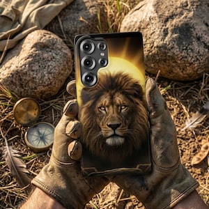 May include: A phone case with a detailed lion's face design, set against a golden sun. The case has a black border and a multi-lens camera system. The background is a natural outdoor scene with rocks and foliage.