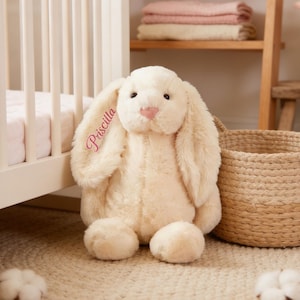 May include: A plush, cream-colored bunny rabbit with long, floppy ears and a pink nose. The name "Priscilla" is embroidered on the ear in pink script. The bunny is sitting on a light-colored rug near a woven basket and a crib.